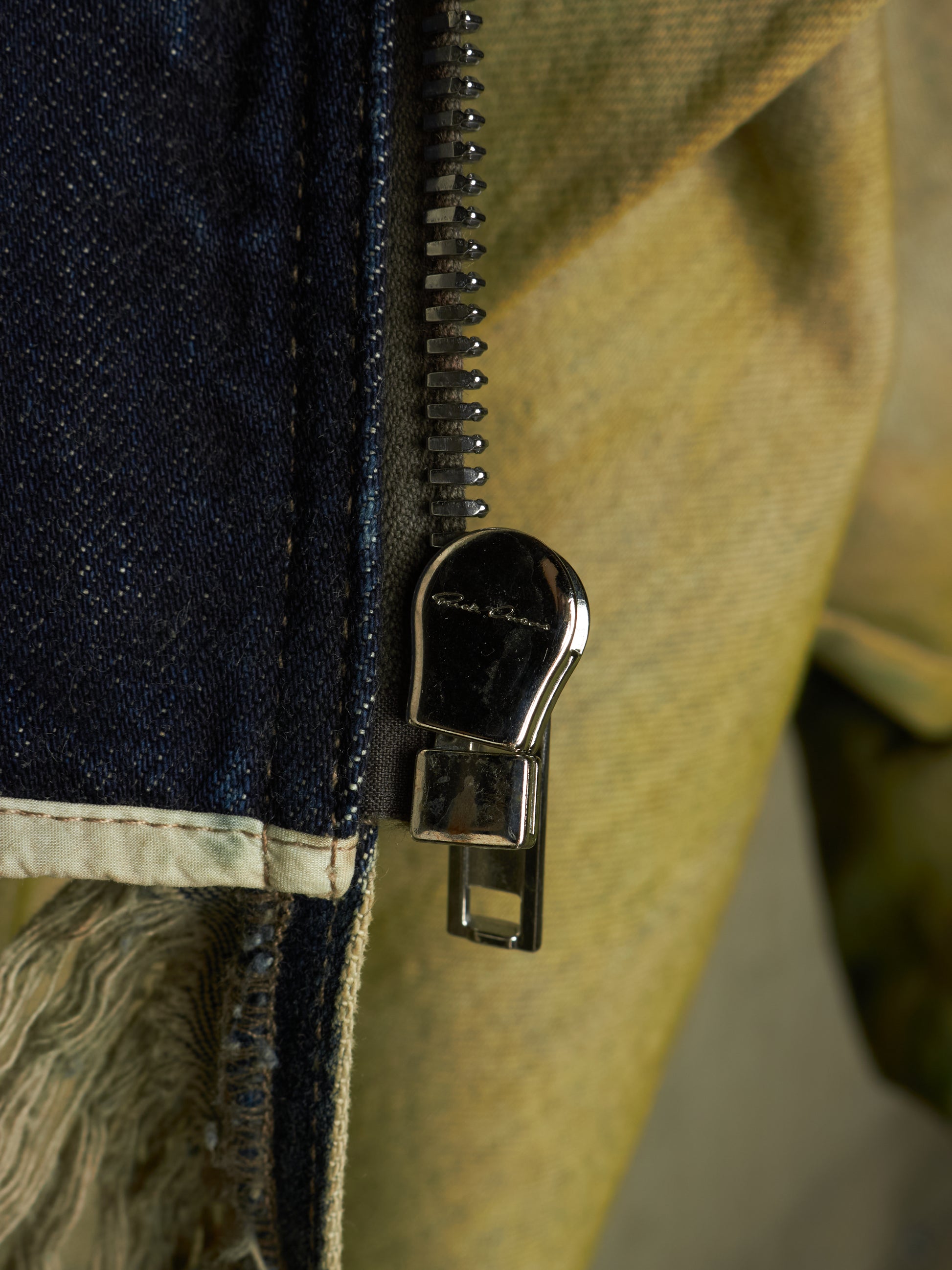 Close-up of a dark metal zipper pull with a small white logo, set between a dark blue denim fabric and a tan canvas material.