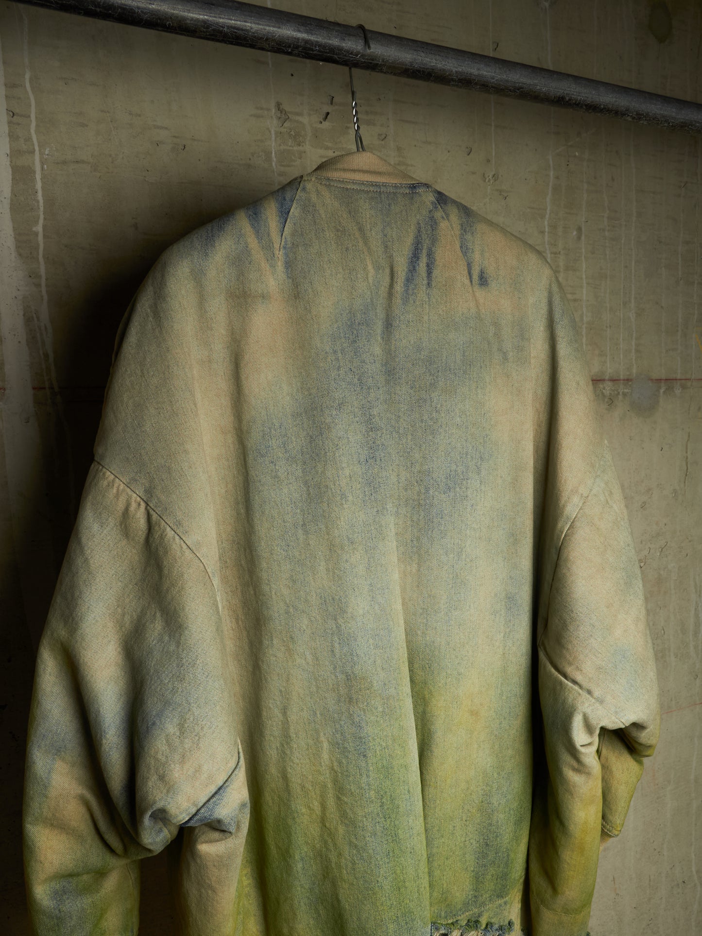 A washed, oversized long-sleeve shirt or jacket hanging on a wooden hanger against a textured concrete wall. The garment features a distressed, faded color palette blending muted yellow-green, olive, and gray-blue tones.