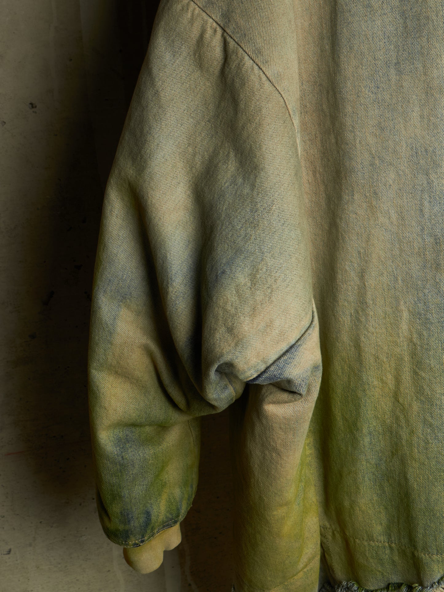 A close-up of a textured, oversized long-sleeve top or jacket in a muted yellow-green color with a distressed, tie-dye-like mottled pattern in olive and gray tones.
