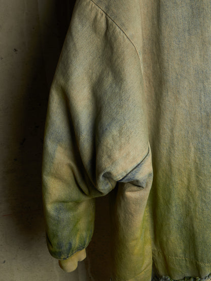 A close-up of a textured, oversized long-sleeve top or jacket in a muted yellow-green color with a distressed, tie-dye-like mottled pattern in olive and gray tones.