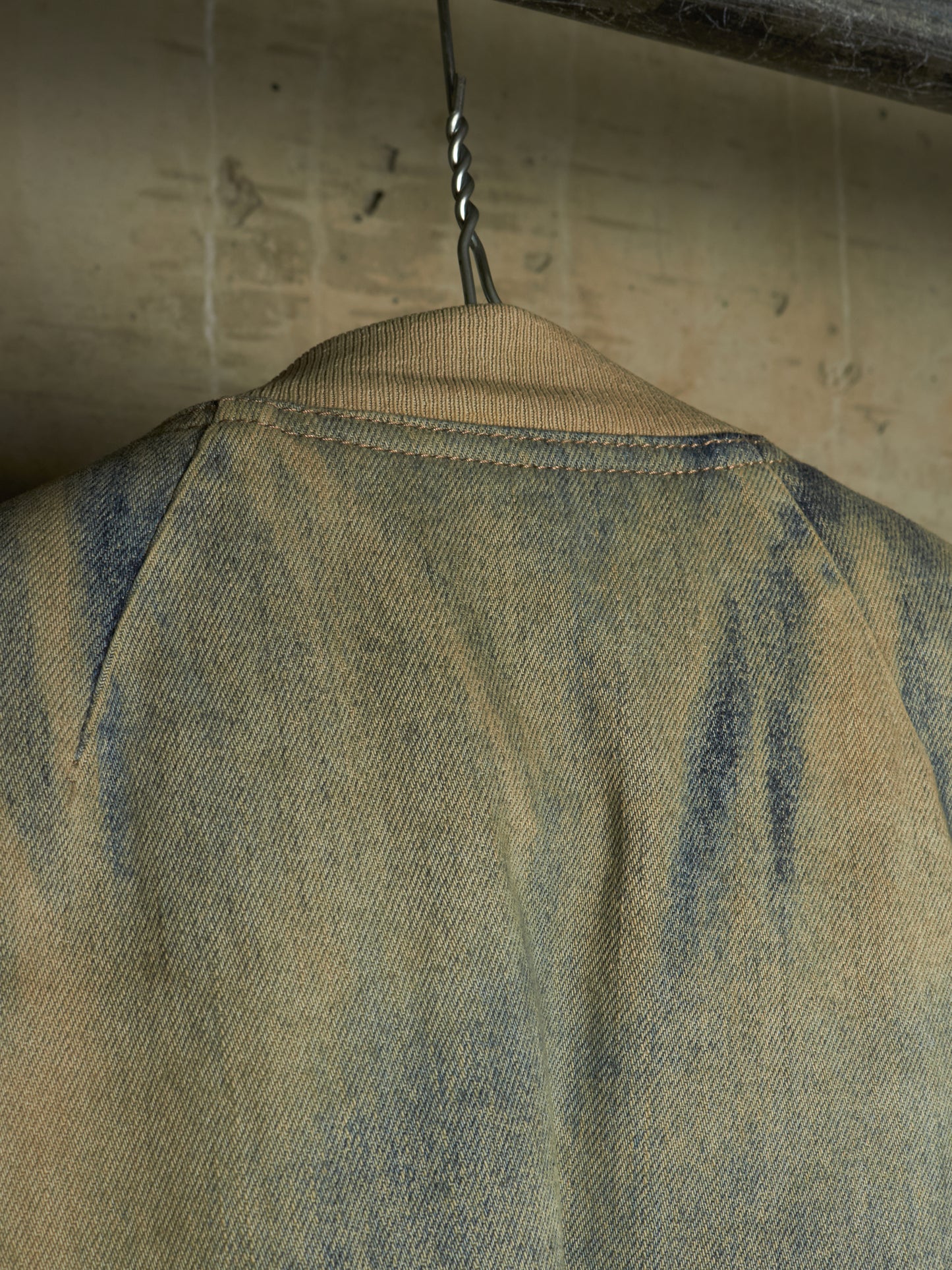 Close-up of the back of a faded blue denim jacket hanging on a metal hook against a textured beige wall.
