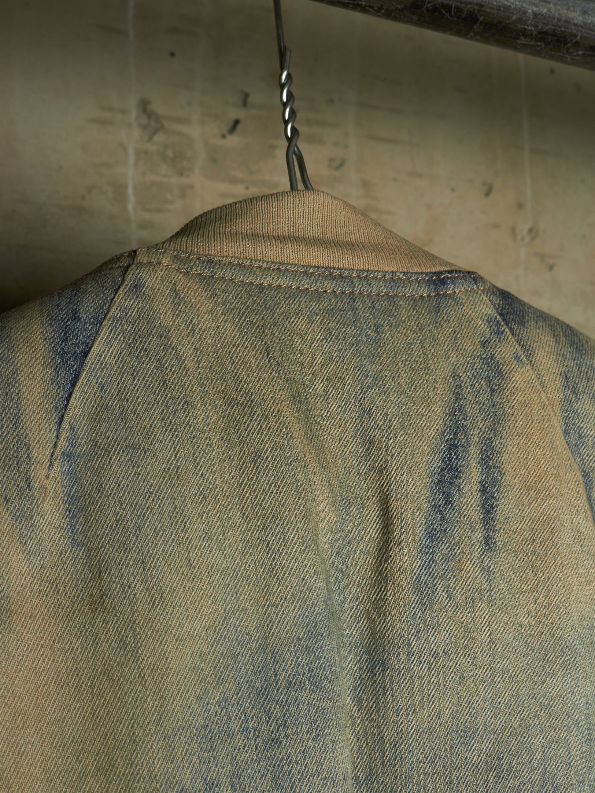 Close-up of the back of a faded blue denim jacket hanging on a metal hook against a textured beige wall.