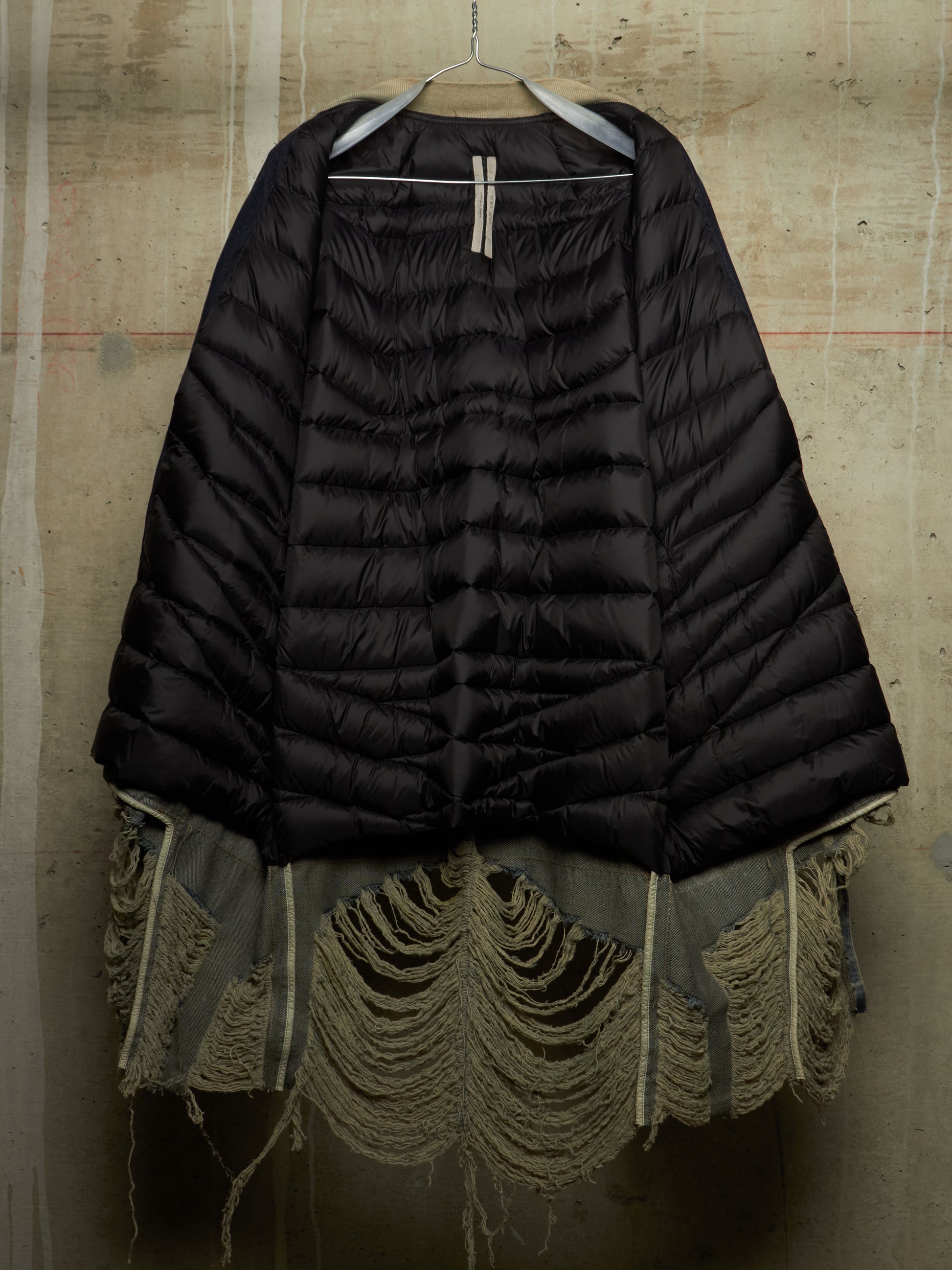A black quilted puffer jacket with a highly asymmetrical, layered hem featuring frayed and tattered olive green fabric layers, hanging against a textured concrete wall.