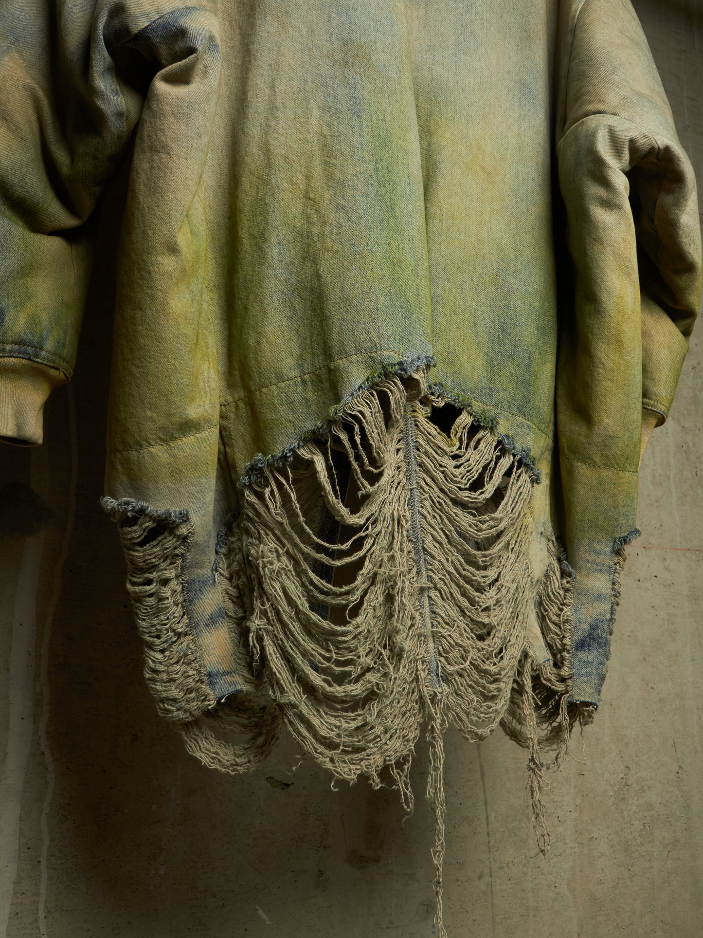 A close-up of a heavily distressed denim jacket with a heavily frayed, shredded hem and cuffs, showing a faded yellow-green and blue wash against a concrete wall.