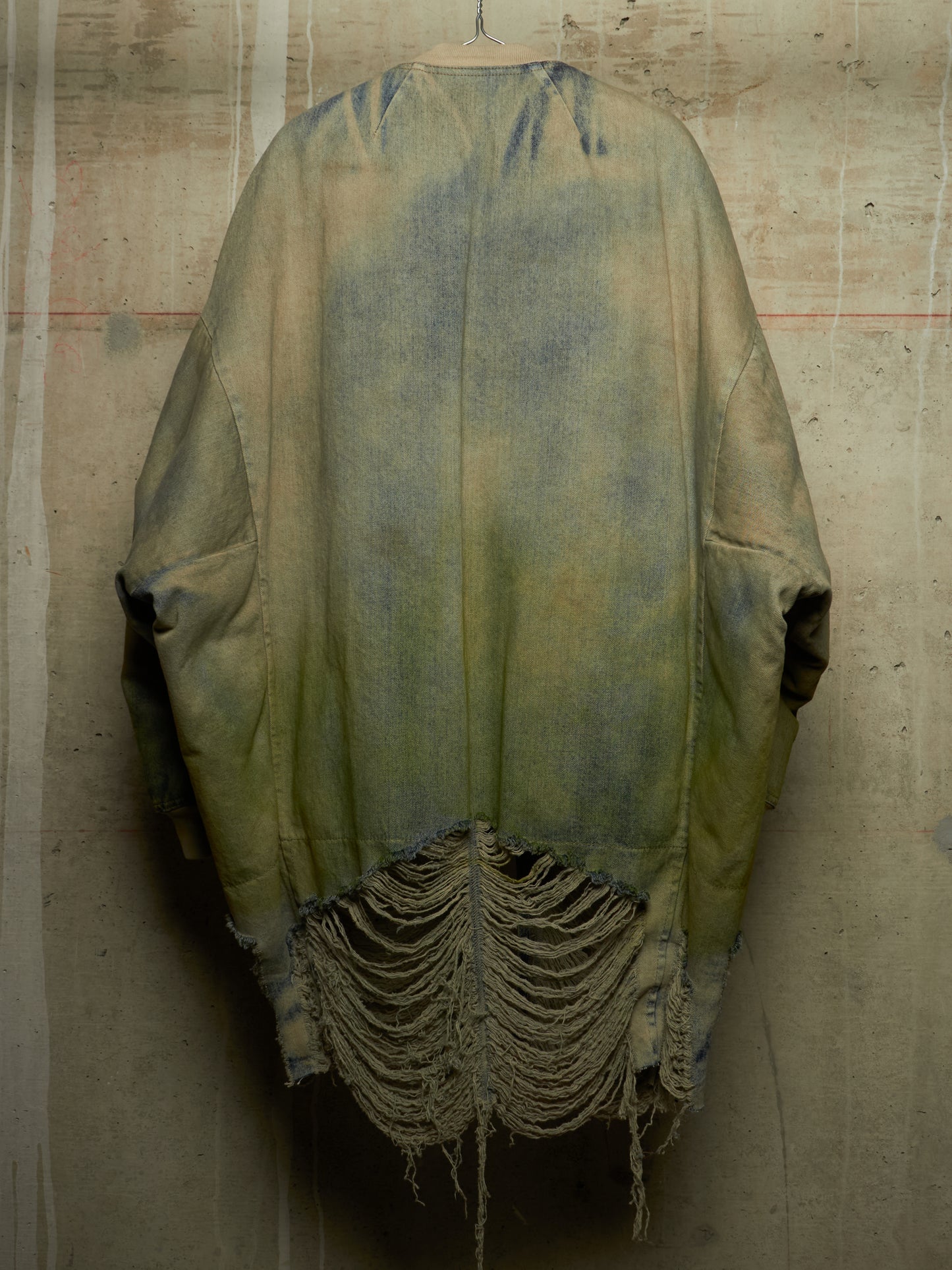 A distressed, oversized poncho-style jacket with a faded olive green and blue wash, hanging against a textured concrete wall. The bottom features heavy frayed, shredded detailing.