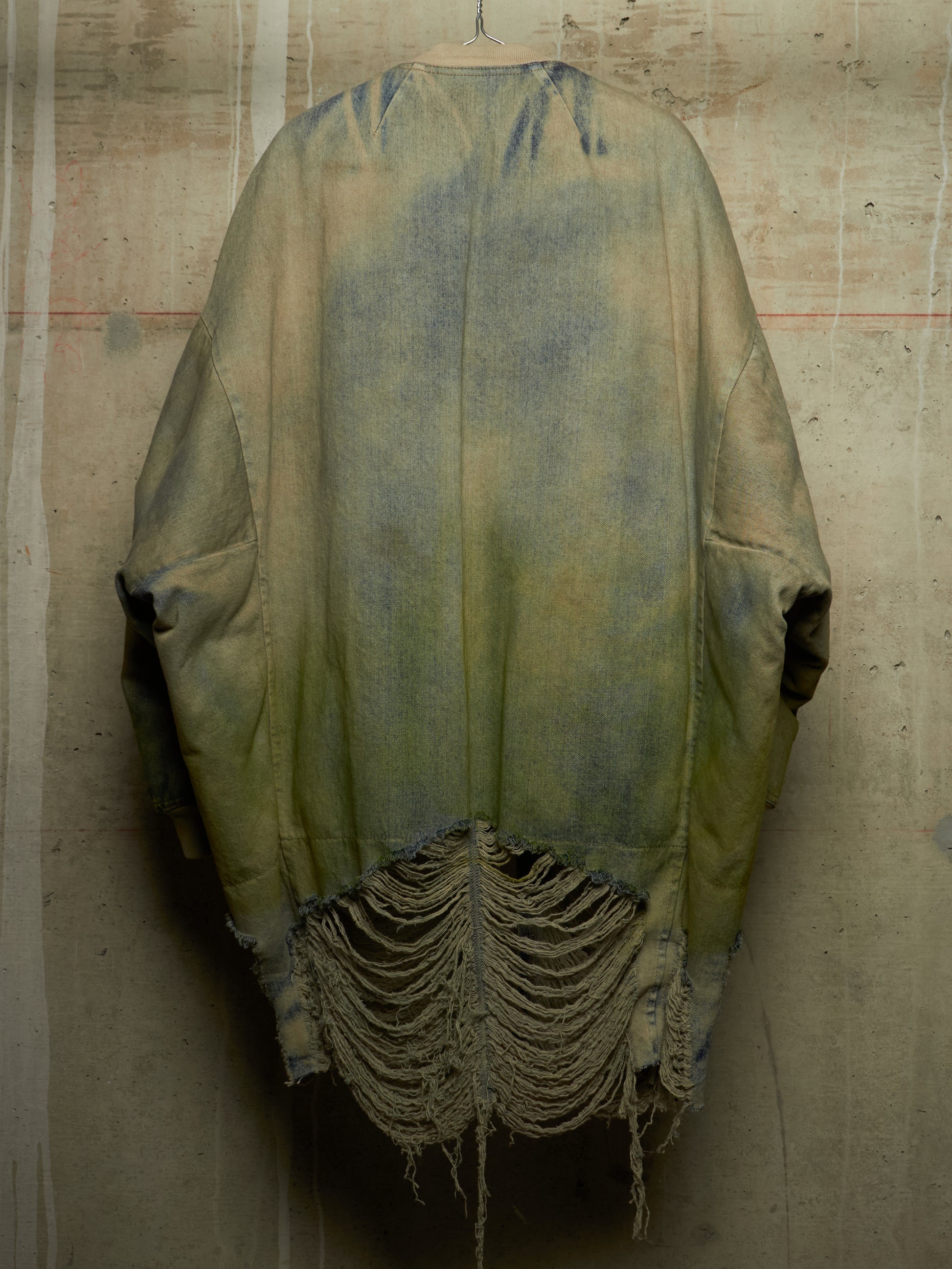 A distressed, oversized poncho-style jacket with a faded olive green and blue wash, hanging against a textured concrete wall. The bottom features heavy frayed, shredded detailing.