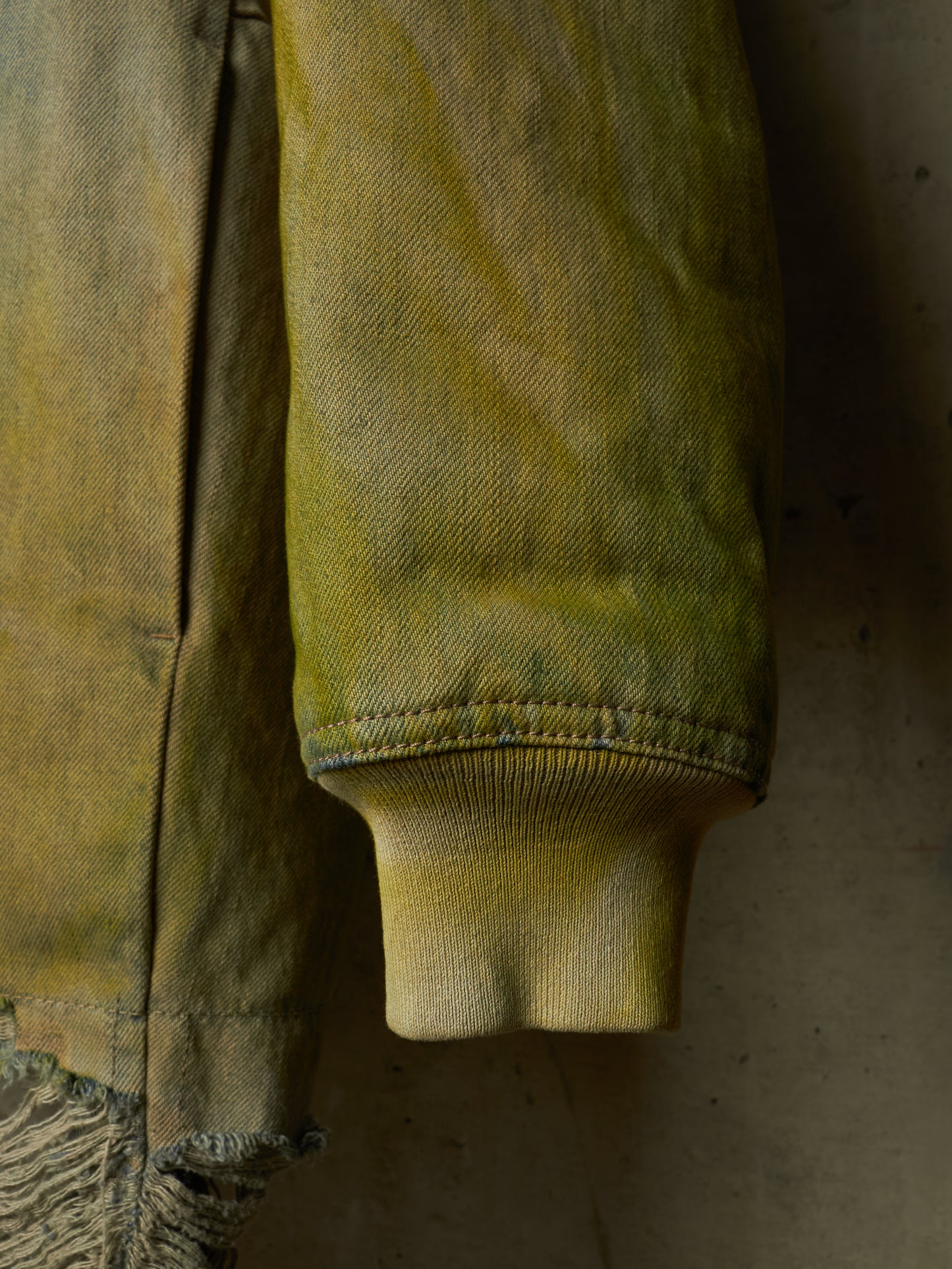 Close-up of a distressed olive green denim jacket sleeve, showing the ribbed cuff and frayed hem against a dark textured background.