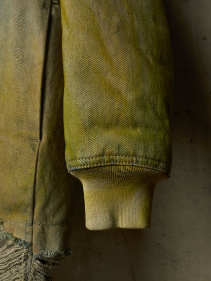 Close-up of a distressed olive green denim jacket sleeve, showing the ribbed cuff and frayed hem against a dark textured background.