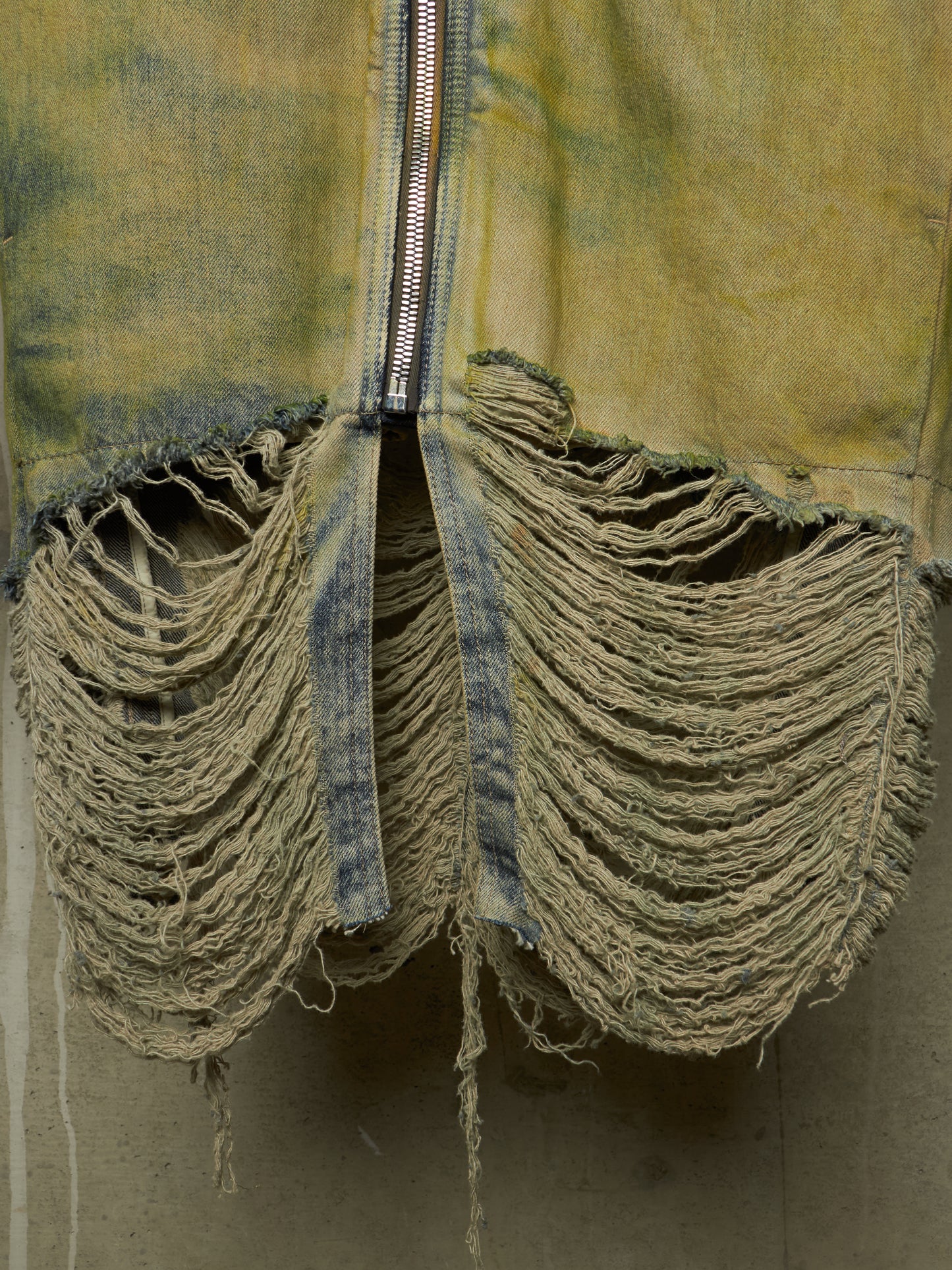 A close-up of a distressed denim garment with heavy fraying and tattered fabric, featuring a central silver zipper.