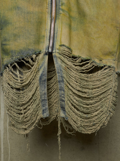 A close-up of a distressed denim garment with heavy fraying and tattered fabric, featuring a central silver zipper.