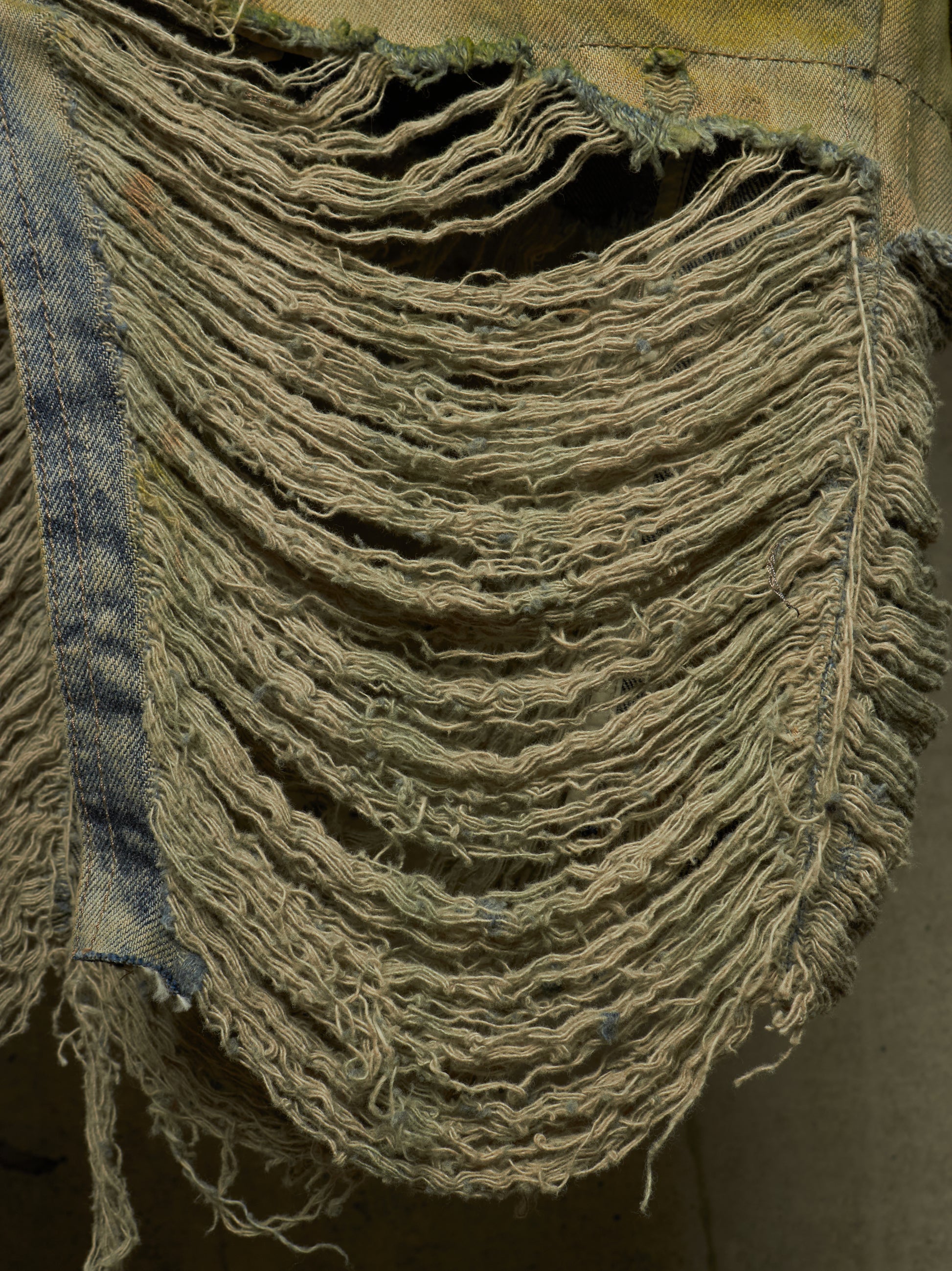 A close-up of a heavily distressed denim garment, showing frayed, shredded layers of light blue and beige denim fabric.