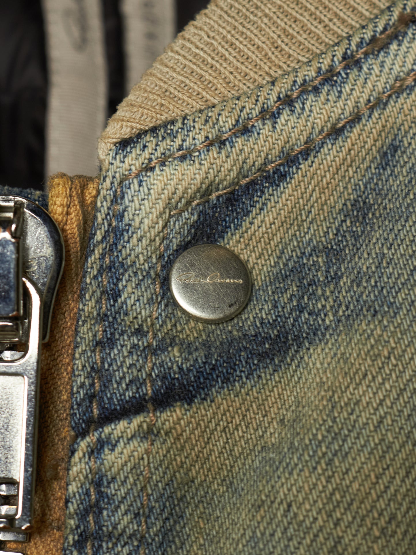 Close-up detail of a vintage denim jacket showing a round silver metal snap button, a silver zipper pull, and a beige ribbed knit collar on a faded blue and tan denim fabric.