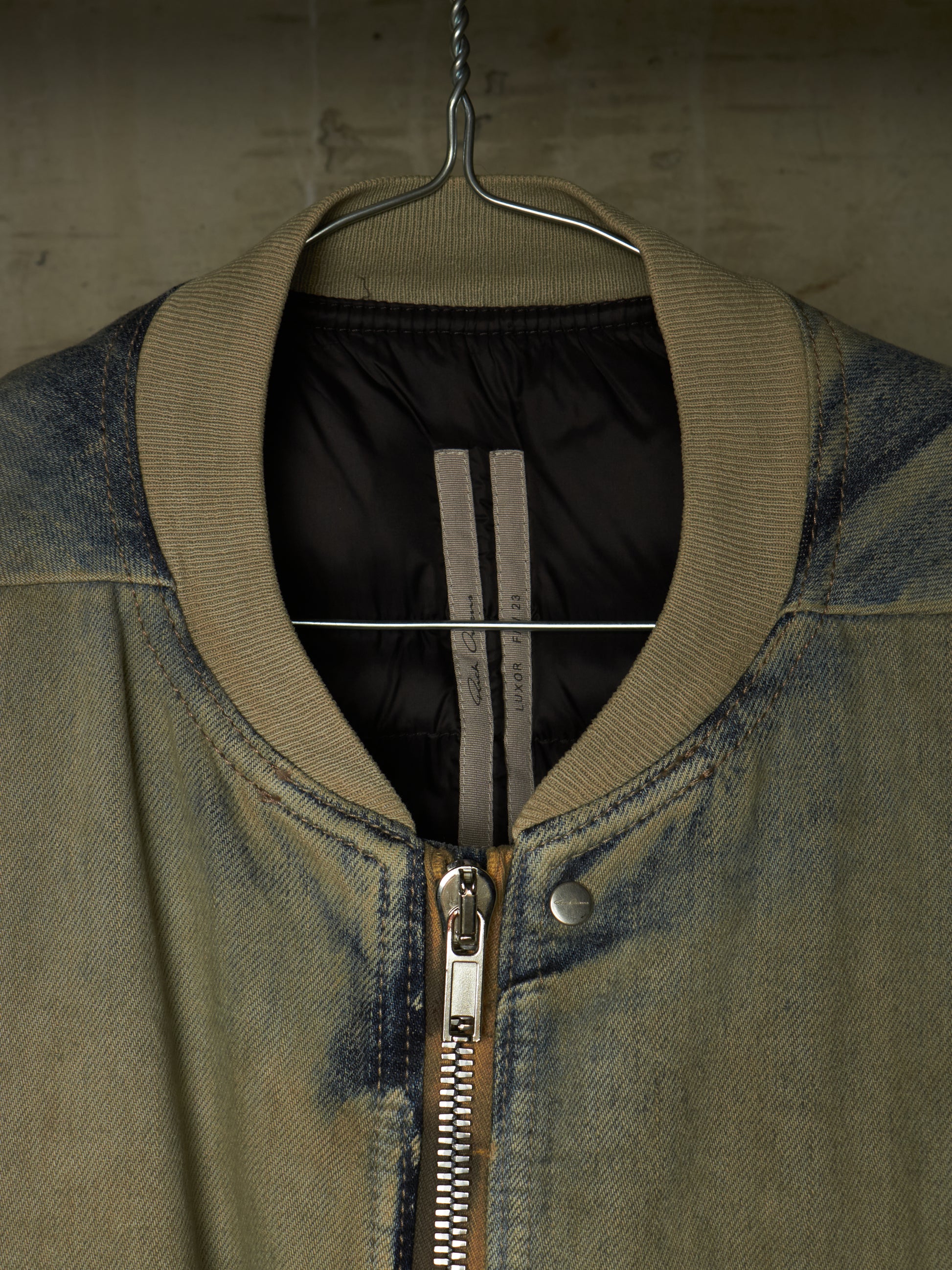 A close-up of a bomber-style jacket hanging on a metal hanger against a textured concrete wall. The jacket has a faded blue-gray denim body, a ribbed olive-khaki collar, a silver metal zipper, and a black quilted interior lining with two vertical gray stripes.