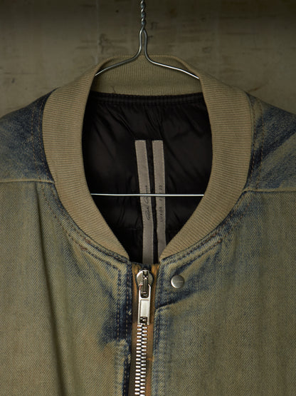 A close-up of a bomber-style jacket hanging on a metal hanger against a textured concrete wall. The jacket has a faded blue-gray denim body, a ribbed olive-khaki collar, a silver metal zipper, and a black quilted interior lining with two vertical gray stripes.