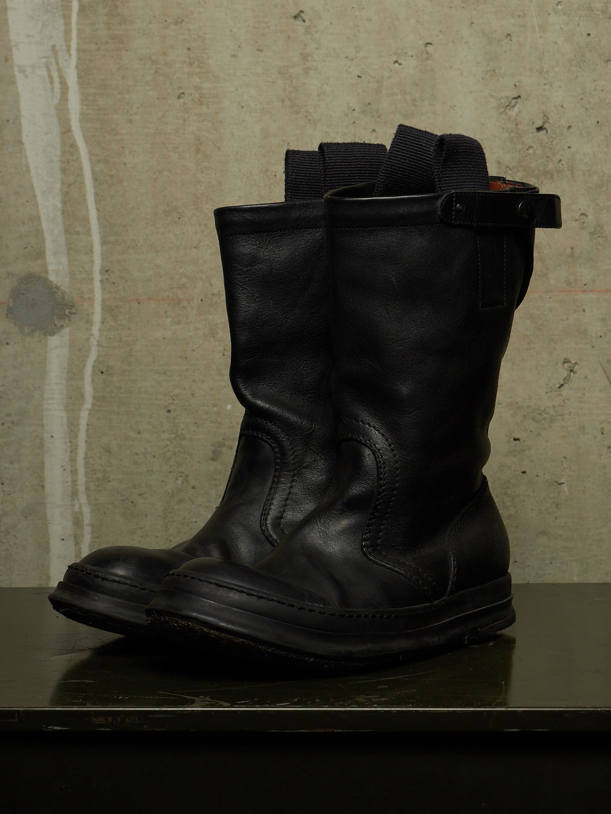 A pair of black leather mid-calf boots with elastic side straps, displayed on a dark surface against a textured concrete wall.