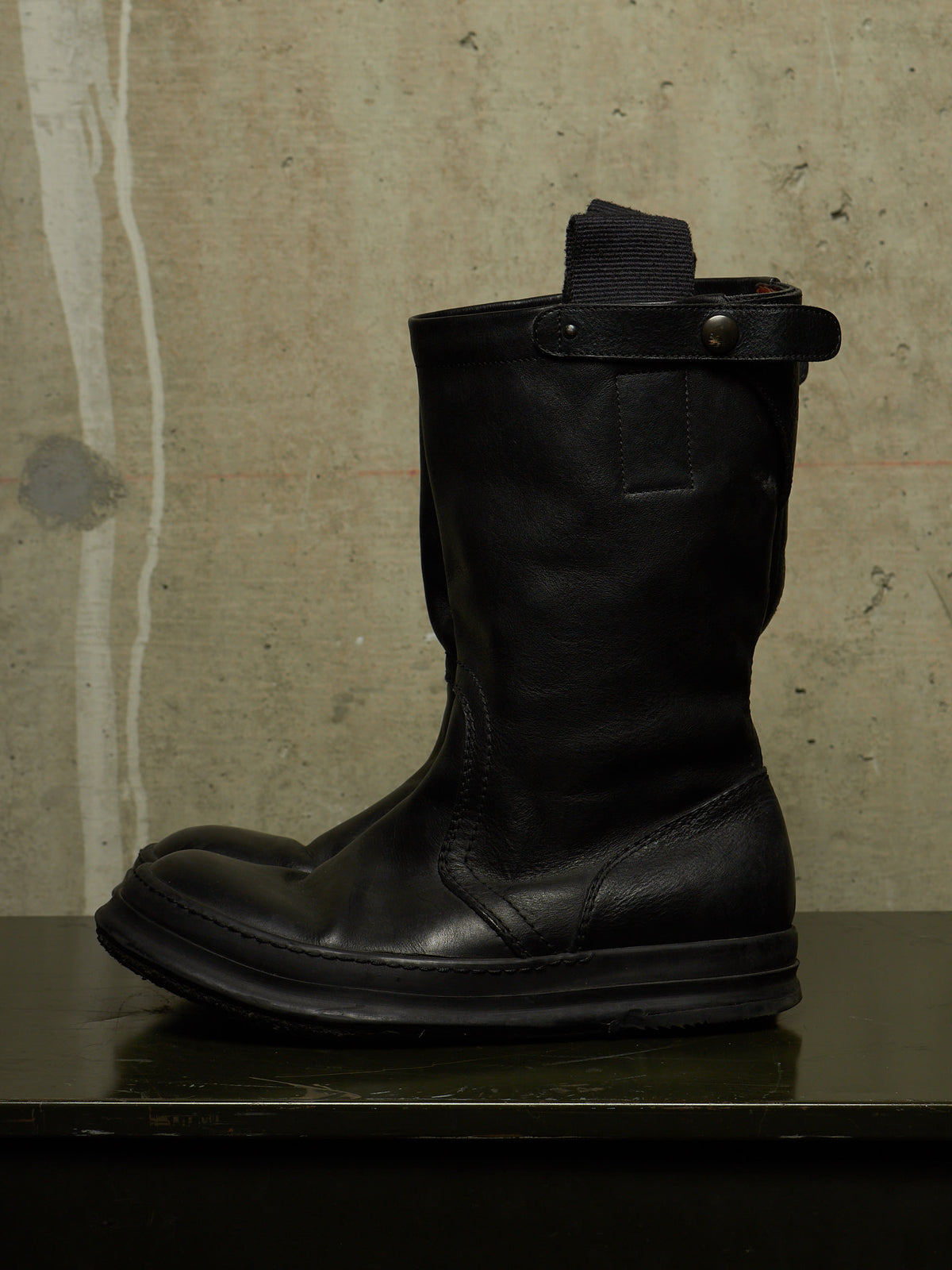 A single black leather mid-calf boot with a strap closure and metal snap, resting on a dark reflective surface against a textured concrete wall.