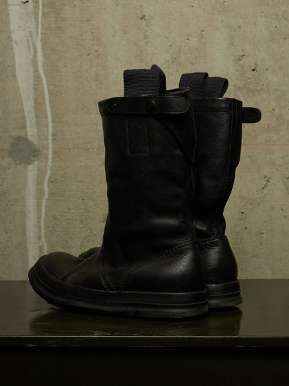 A pair of black leather mid-calf boots with elastic side panels and pull tabs, standing on a dark surface against a textured concrete wall.