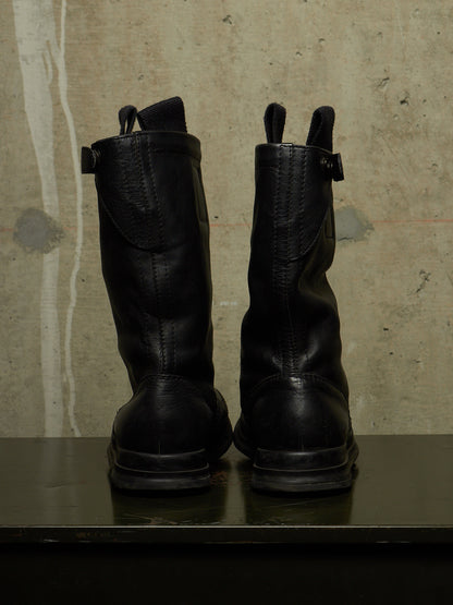 A pair of black leather mid-calf boots shown from the back, standing on a dark surface against a textured concrete wall.