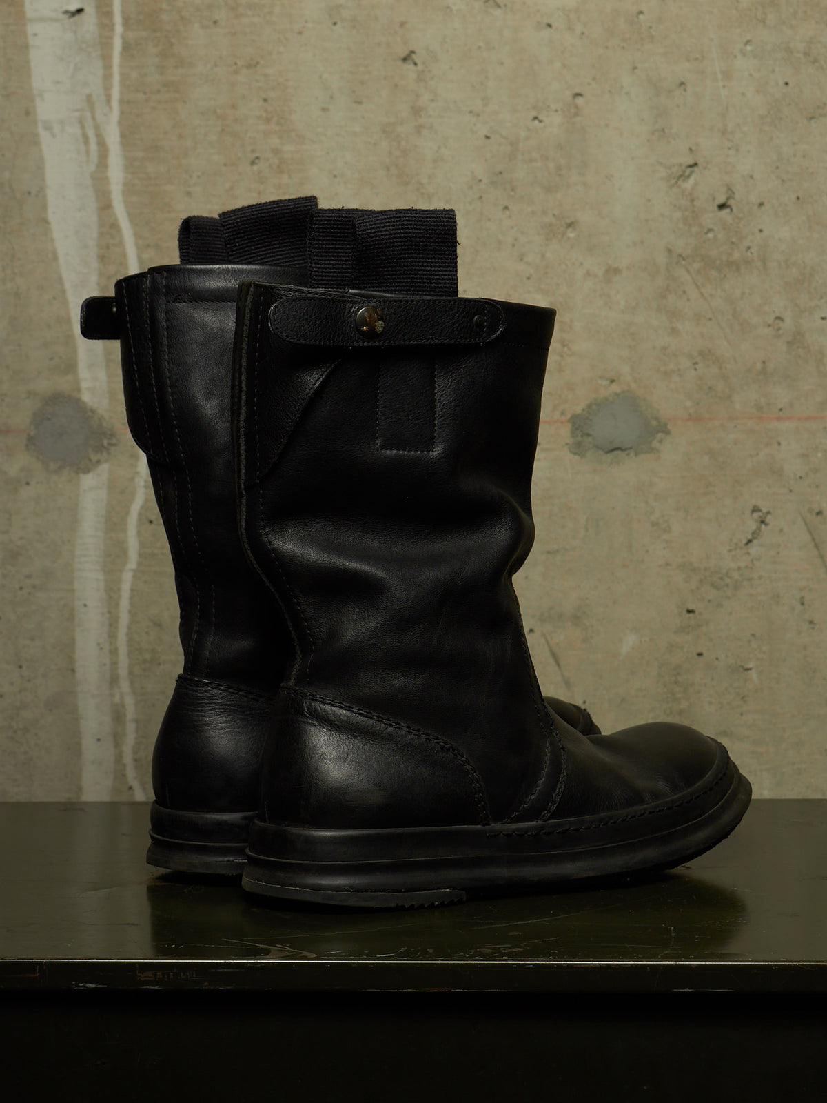 A pair of black leather mid-calf boots with elastic side panels and a snap tab detail, displayed on a dark surface against a textured concrete wall.