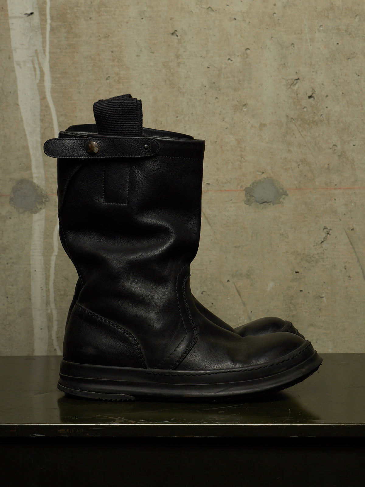 A single black leather mid-calf boot with a pull tab and strap detail, displayed on a dark surface against a textured concrete wall.