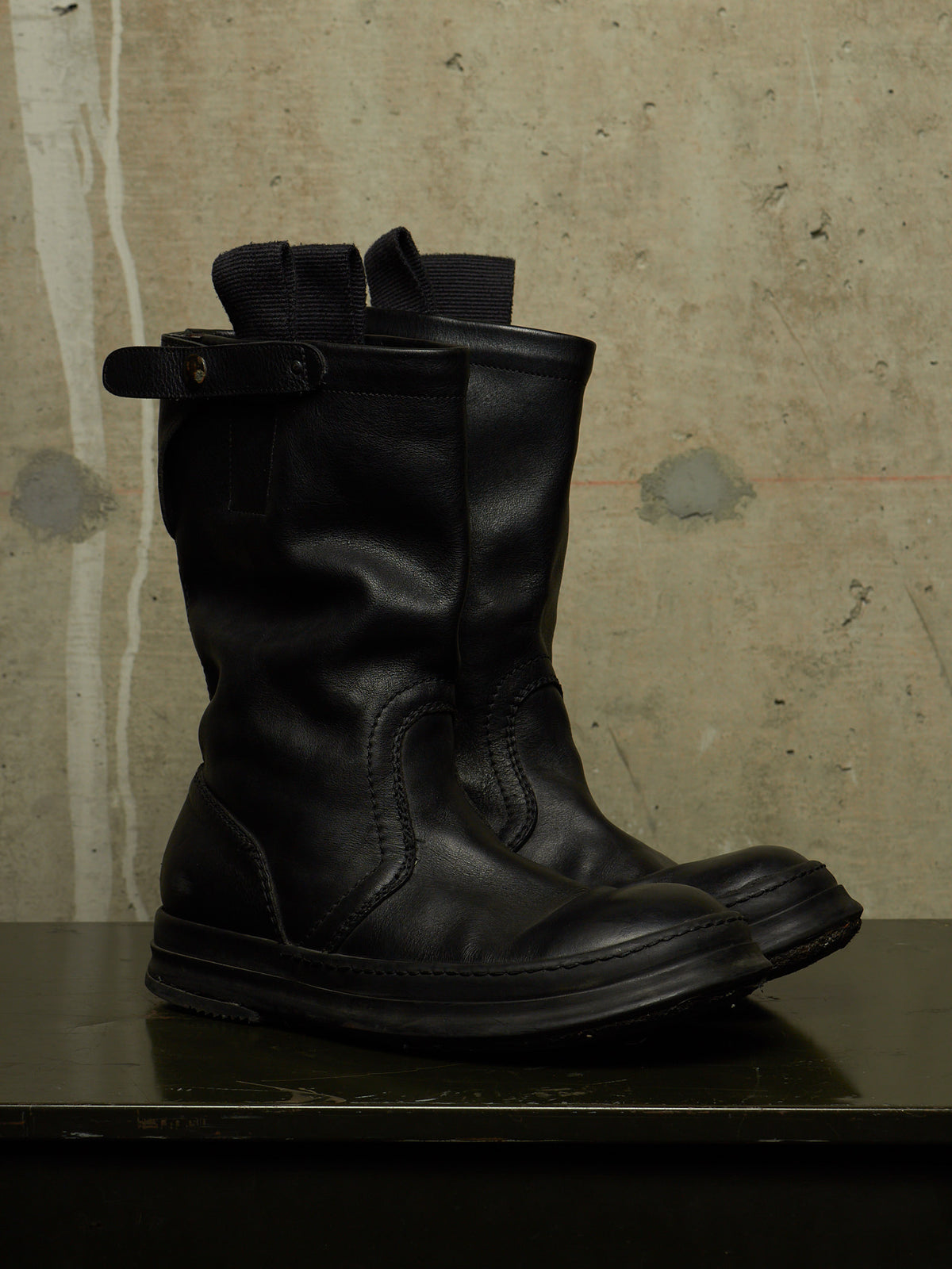 A pair of black leather mid-calf boots with a buckle strap detail on the back, displayed on a dark surface against a textured concrete wall.