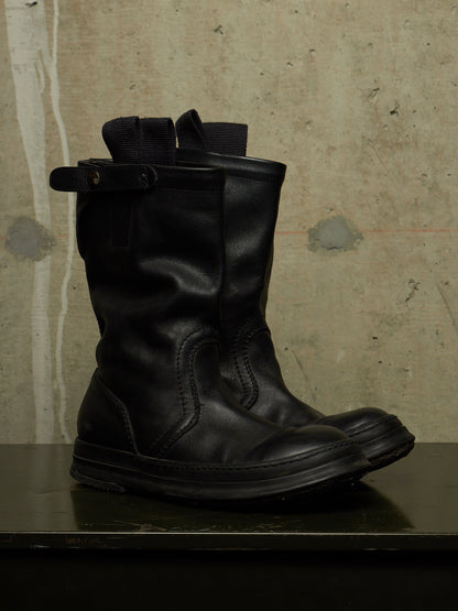 A pair of black leather mid-calf boots with a buckle strap detail on the back, displayed on a dark surface against a textured concrete wall.