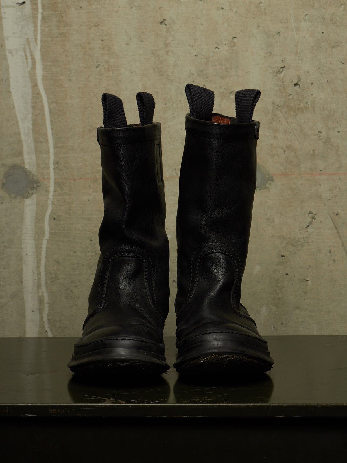 A pair of black leather mid-calf boots with pull tabs, standing side-by-side on a dark reflective surface against a textured concrete wall.