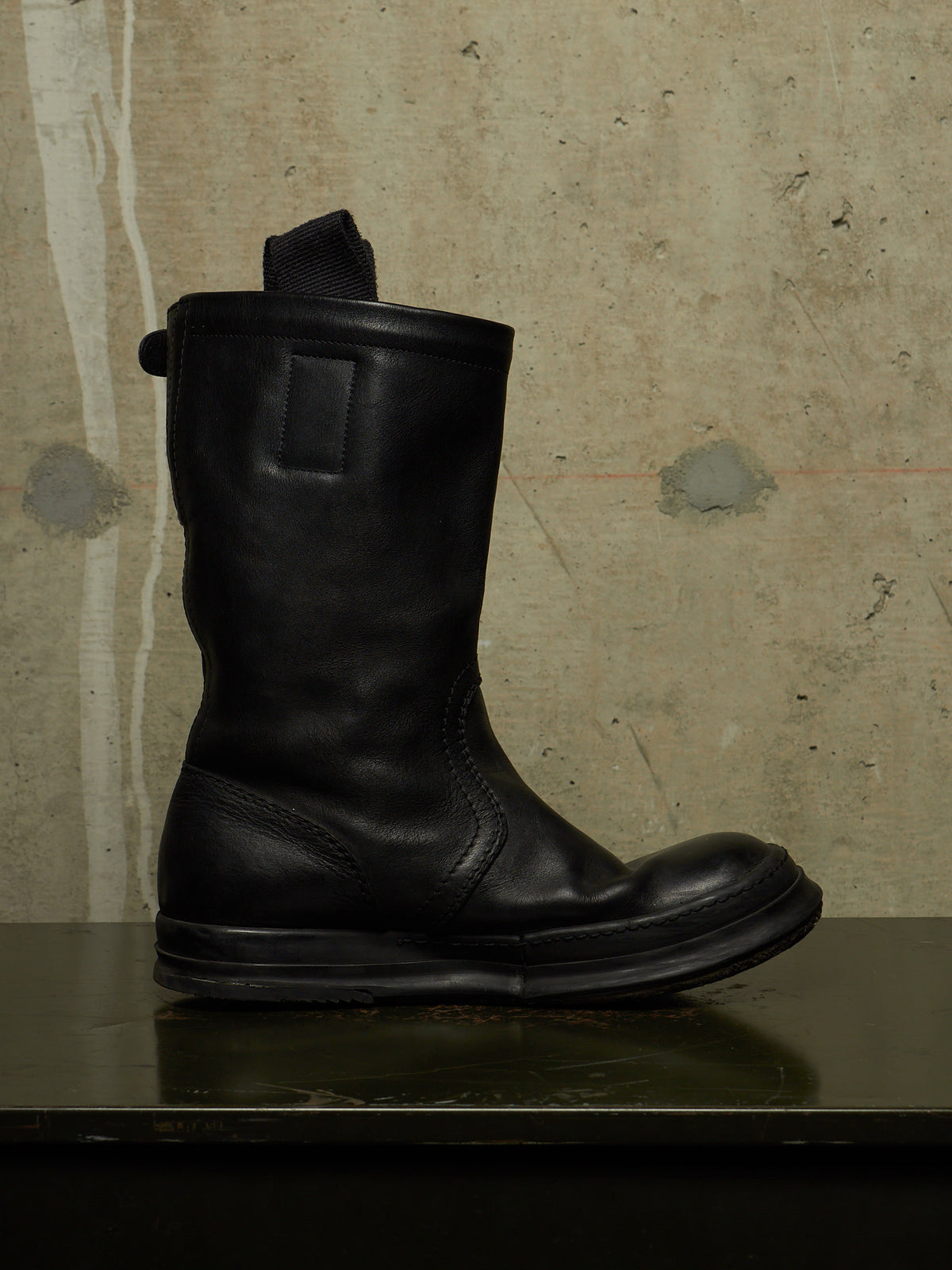 A single black leather mid-calf boot with a pull tab, positioned on a dark reflective surface against a textured concrete wall.