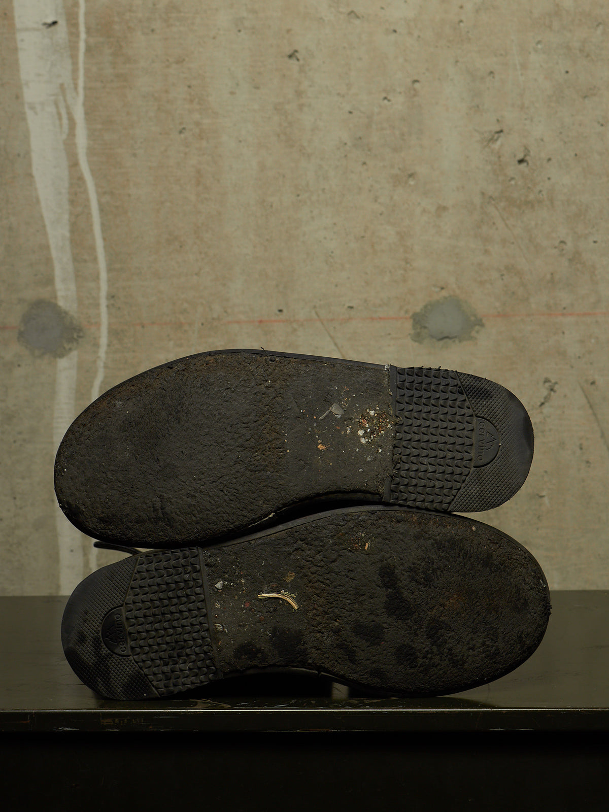 A pair of worn black shoes shown from the sole side, resting on a dark surface against a textured concrete wall.