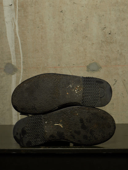 A pair of worn black shoes shown from the sole side, resting on a dark surface against a textured concrete wall.