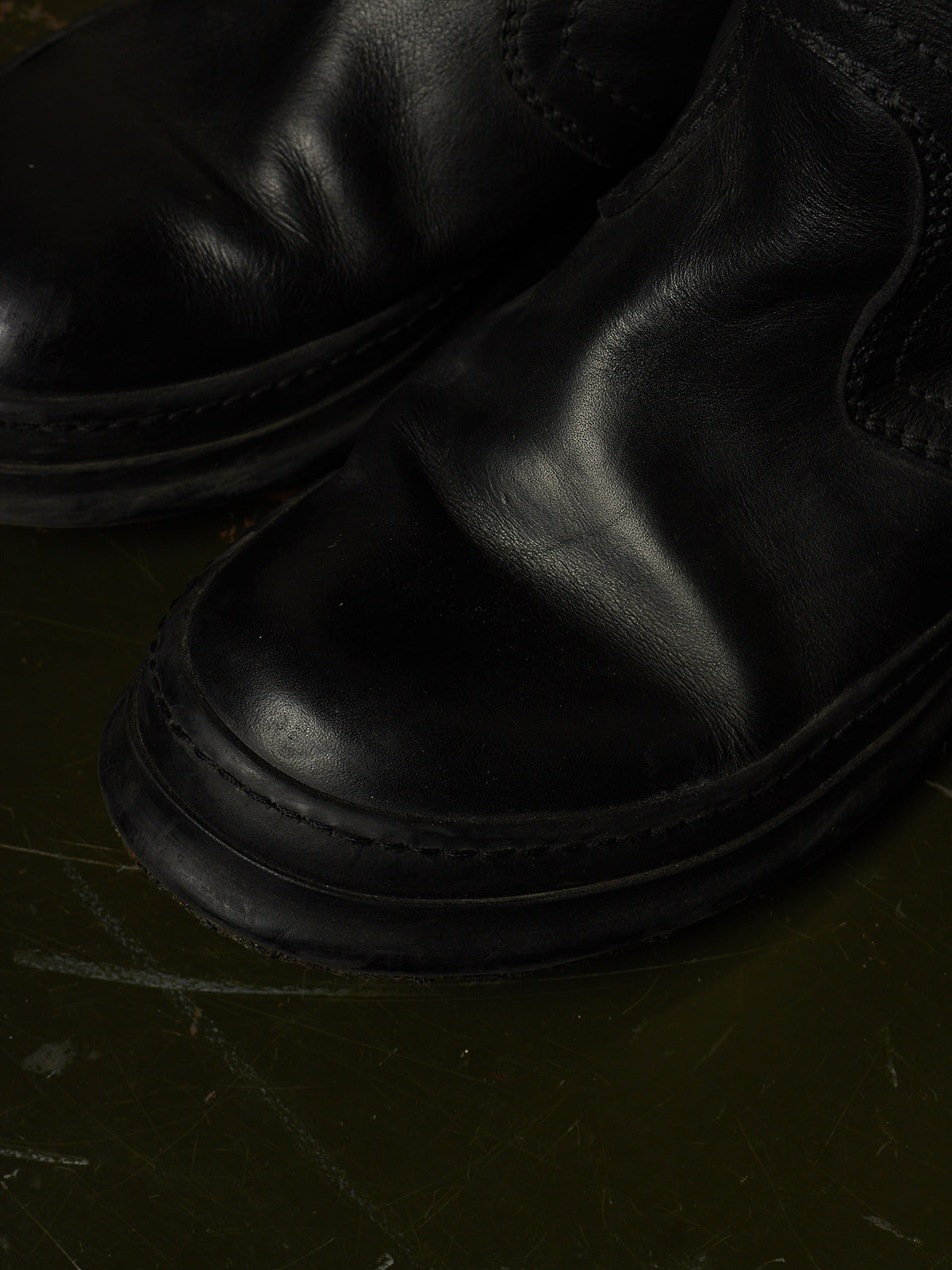 A close-up shot of a pair of black leather boots, highlighting the smooth texture and stitching on a dark, textured surface.
