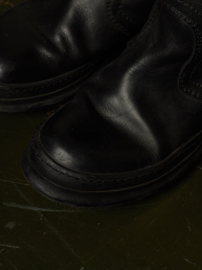 A close-up shot of a pair of black leather boots, highlighting the smooth texture and stitching on a dark, textured surface.