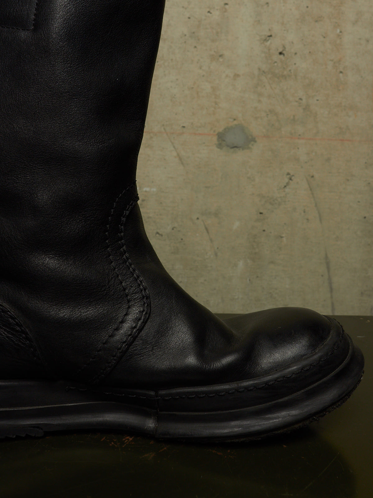 A close-up side view of a black leather boot with a thick black sole, positioned against a textured concrete wall background.