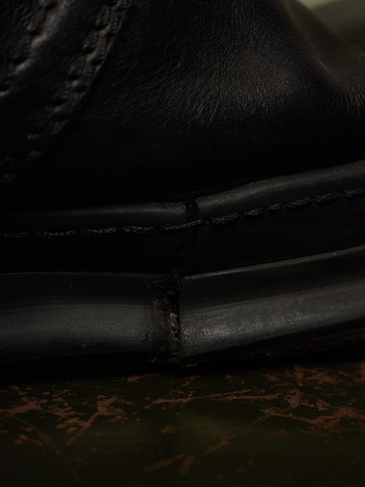 Close-up detail shot of a black leather shoe showing the stitching and sole construction, resting on a dark textured surface.
