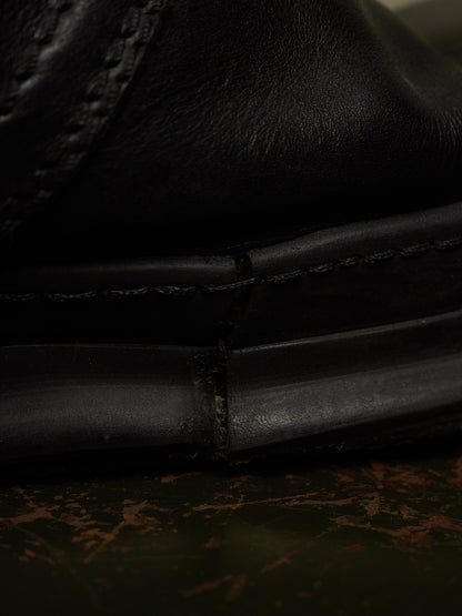 Close-up detail shot of a black leather shoe showing the stitching and sole construction, resting on a dark textured surface.