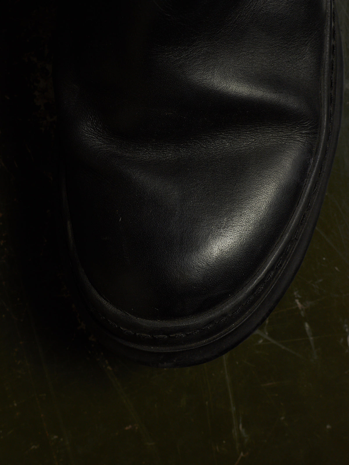 Close-up of a black leather boot toe showing the smooth leather texture and stitched edge against a dark olive-green floor.