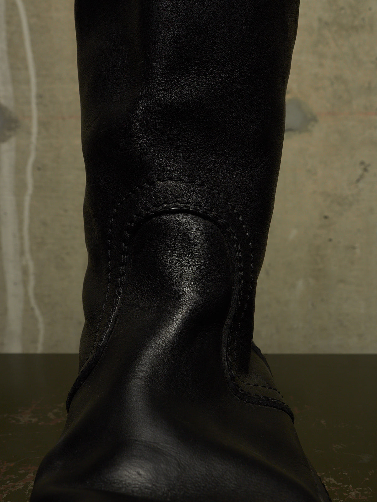 A close-up of a black leather boot, showing the textured leather and decorative stitching against a neutral concrete background.