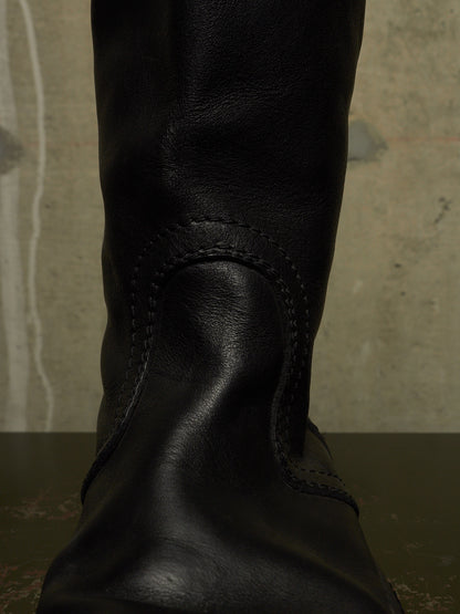A close-up of a black leather boot, showing the textured leather and decorative stitching against a neutral concrete background.