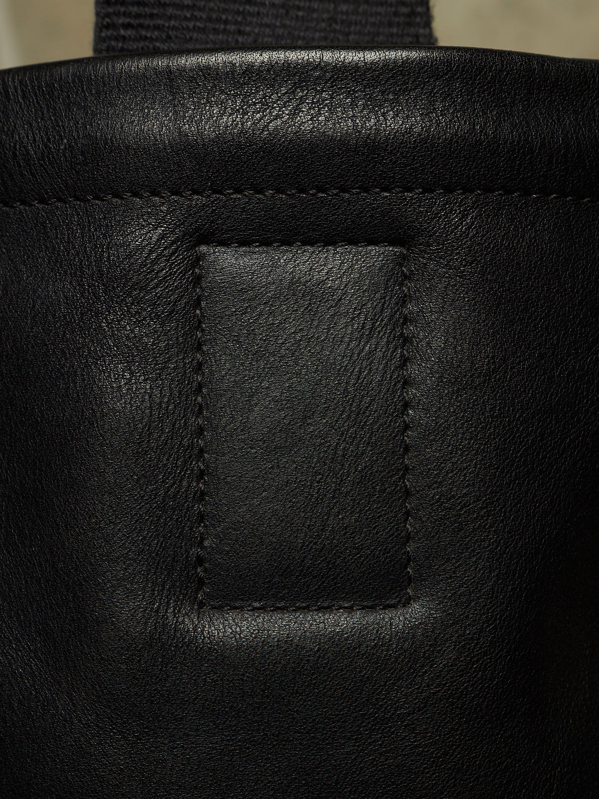 Close-up detail of a black leather bag showing a stitched rectangular patch and part of a dark strap.