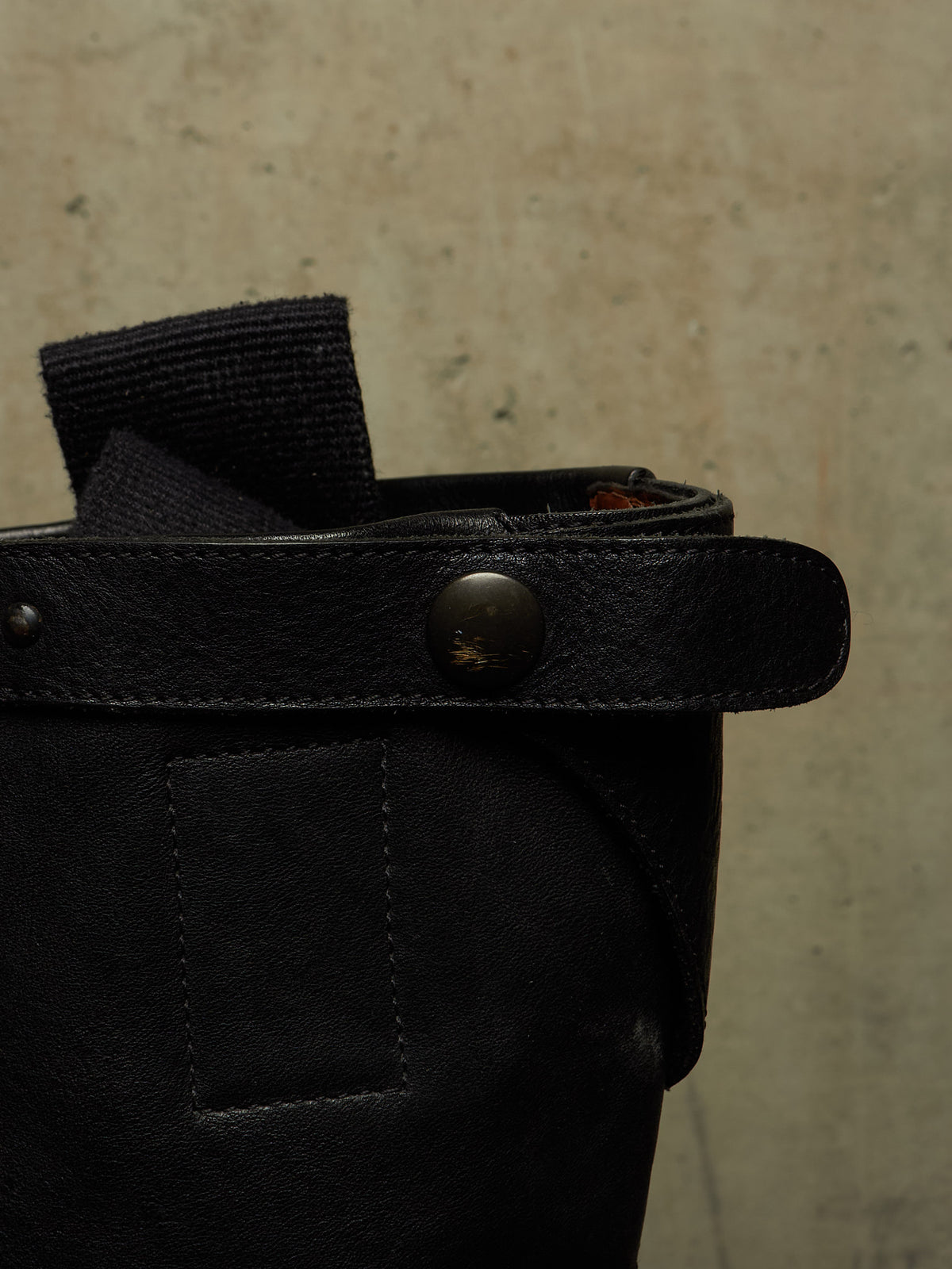 Close-up of a black leather and canvas bag strap with a metal snap button, set against a textured beige concrete wall.