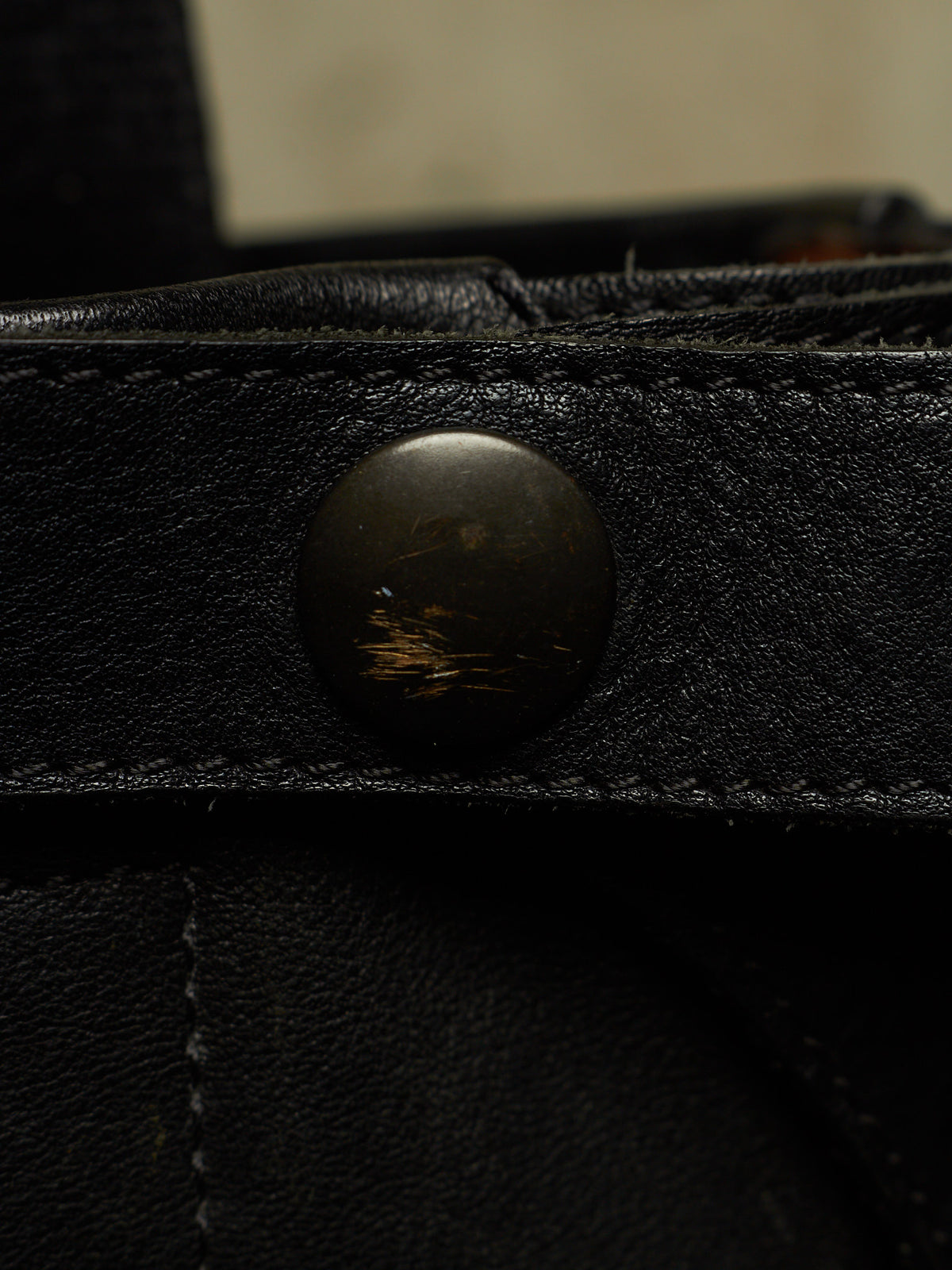 Close-up of a black leather strap with a round dark metal snap button, showing the textured leather surface and stitching.