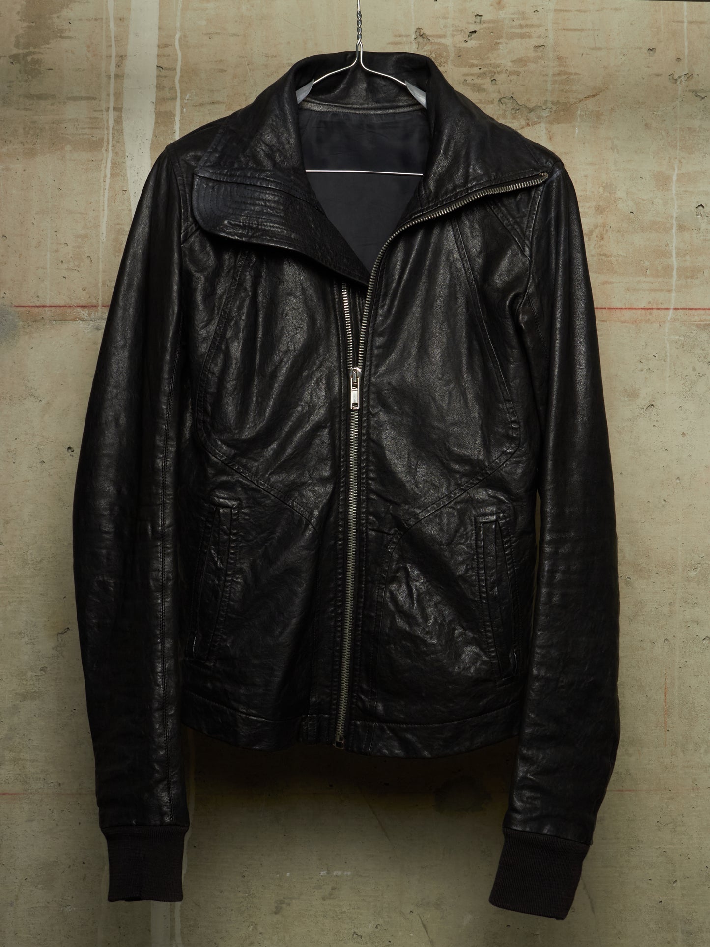 A black leather jacket hanging on a metal hanger against a textured concrete wall. The jacket features a high collar, a front zipper closure, and ribbed cuffs and hem.