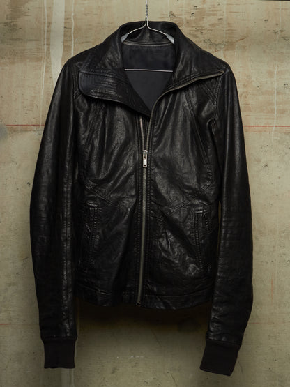 A black leather jacket hanging on a metal hanger against a textured concrete wall. The jacket features a high collar, a front zipper closure, and ribbed cuffs and hem.