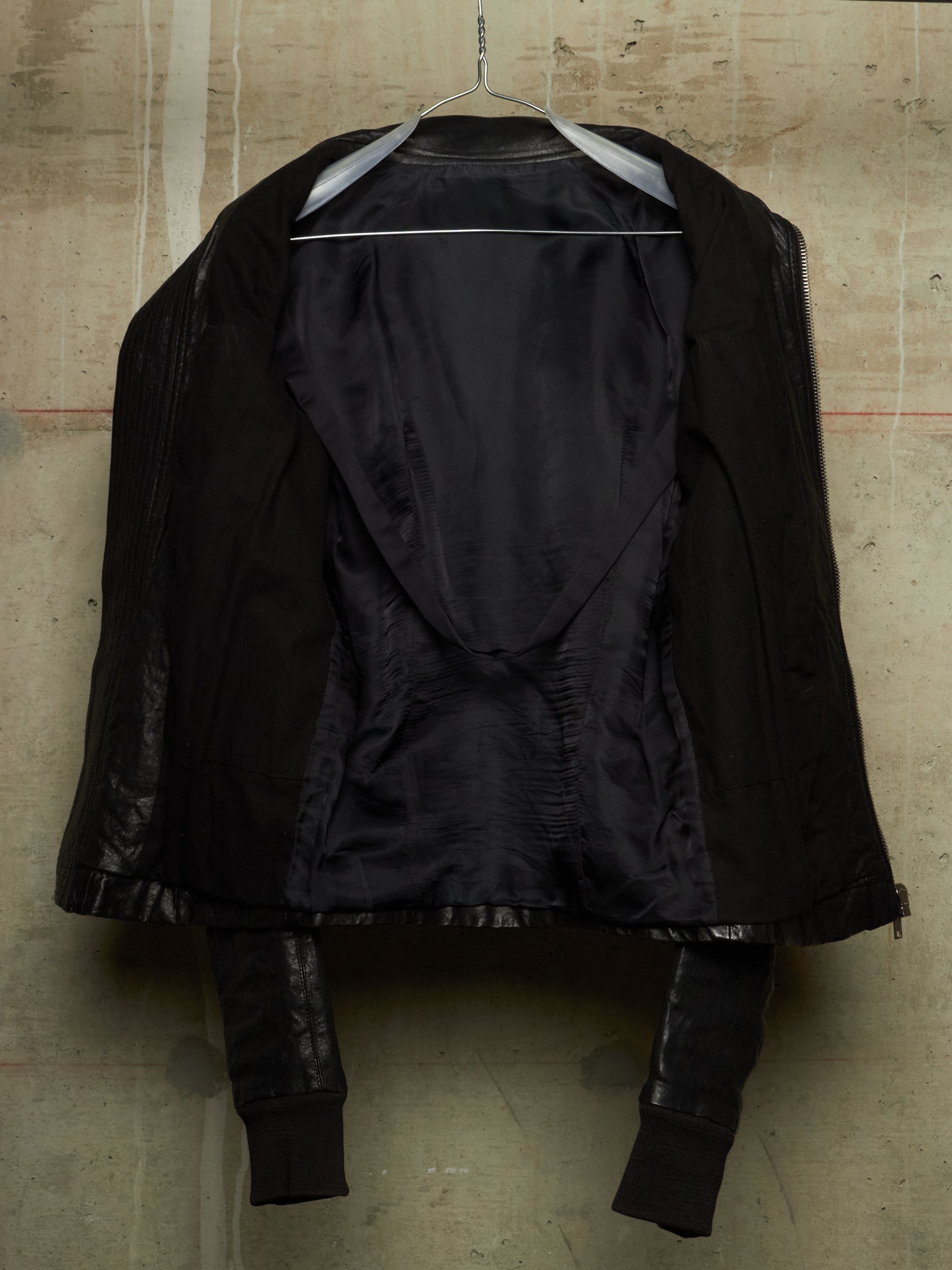 A black structured jacket with a dramatic asymmetrical draped front, hanging on a wire hanger against a textured concrete wall.