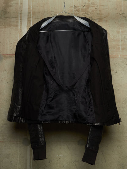 A black structured jacket with a dramatic asymmetrical draped front, hanging on a wire hanger against a textured concrete wall.