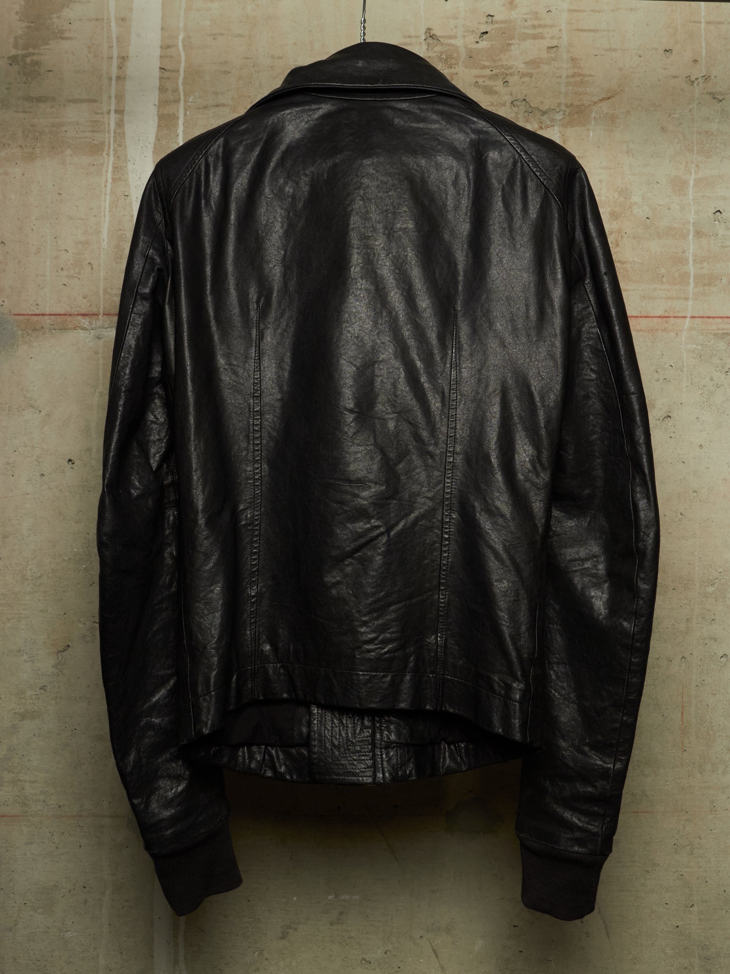 A black leather jacket shown from the back, hanging against a textured concrete wall.