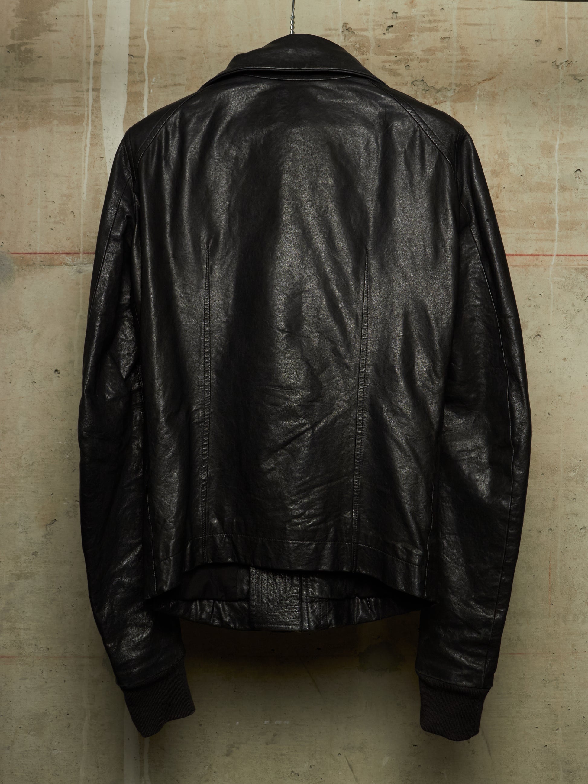 A black leather jacket shown from the back, hanging against a textured concrete wall.