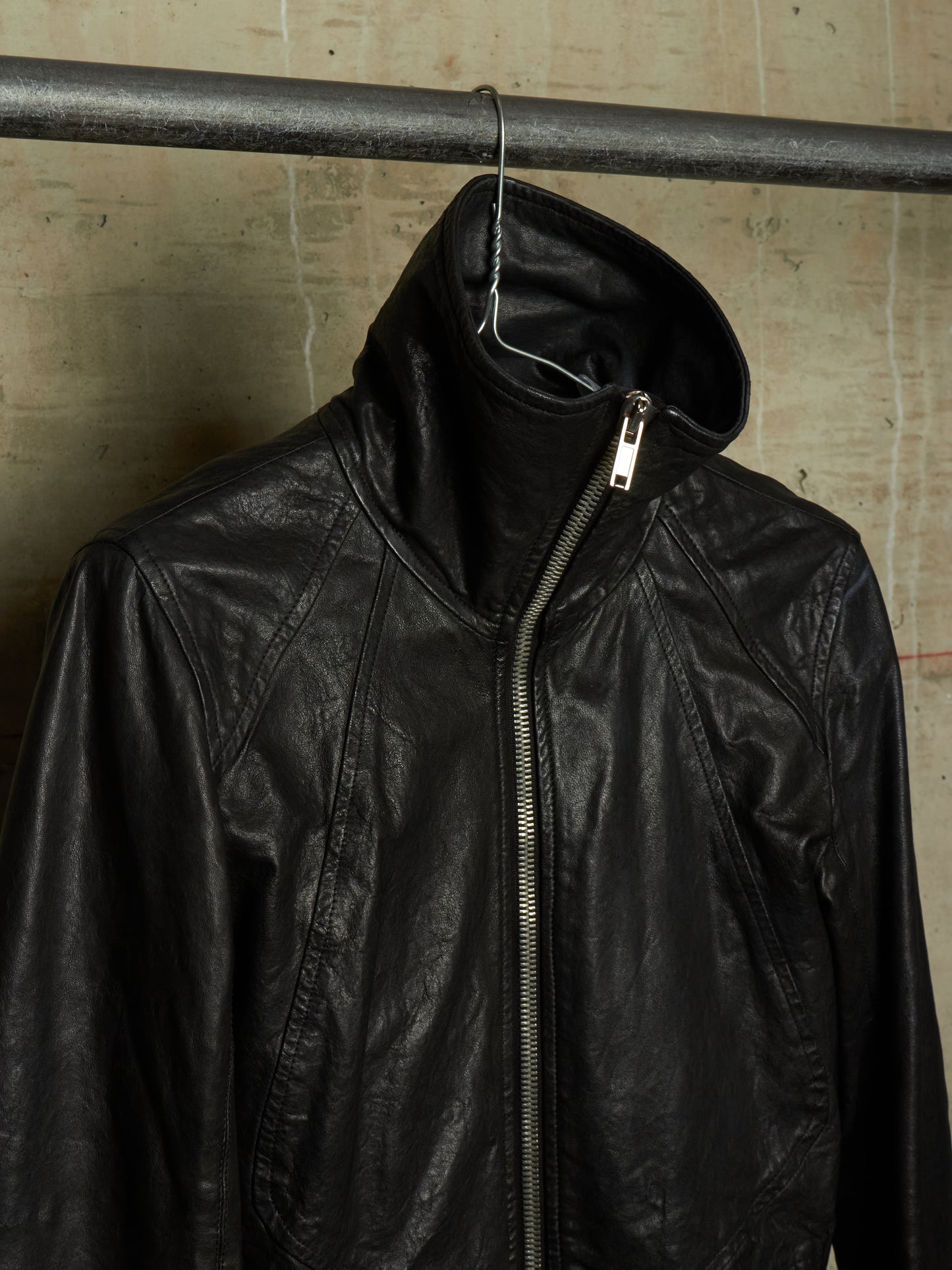 A black leather jacket with a high collar and front zipper, hanging on a metal rod against a textured concrete wall.