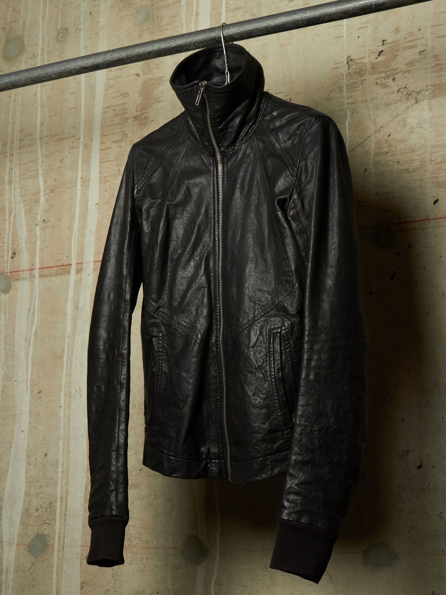 A black leather jacket with a high collar and front zipper, hanging from a metal rod against a textured concrete wall.