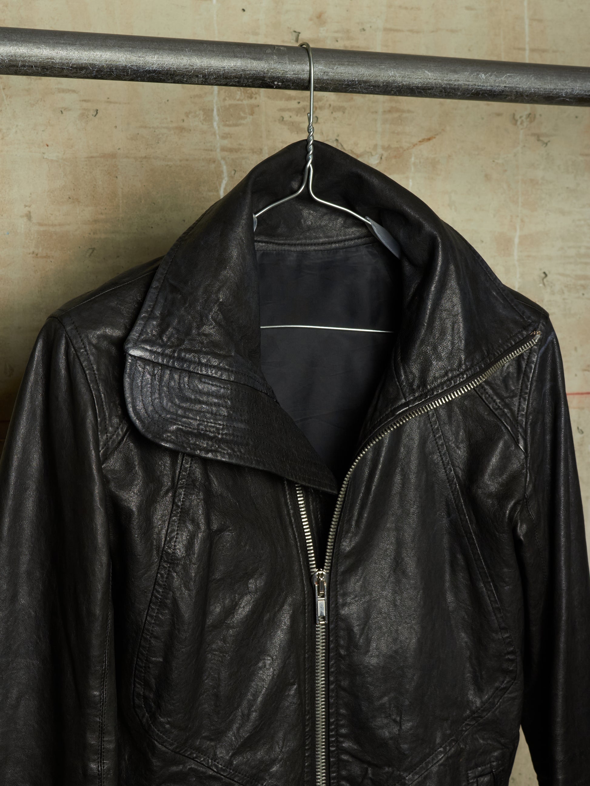 A black leather motorcycle jacket hanging on a metal hanger from a horizontal industrial pipe, set against a textured beige concrete wall.